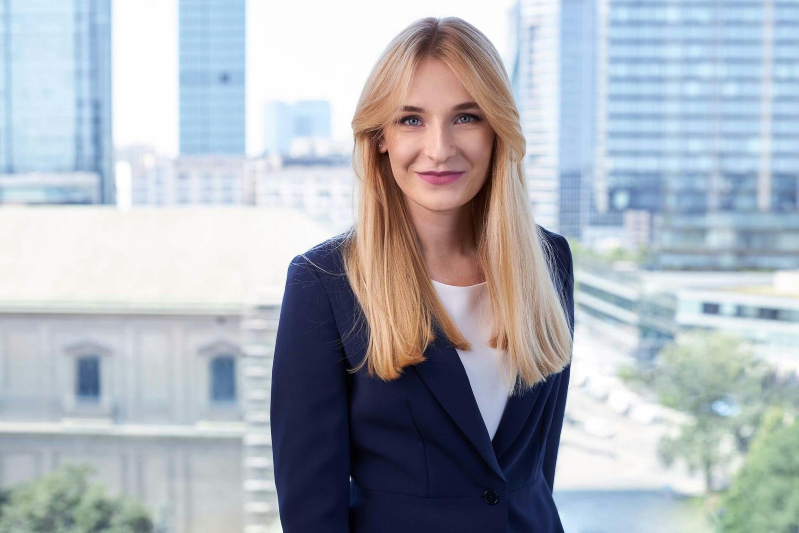 Smiling blonde businesswoman in elegant attire standing against a Warsaw urban backdrop.